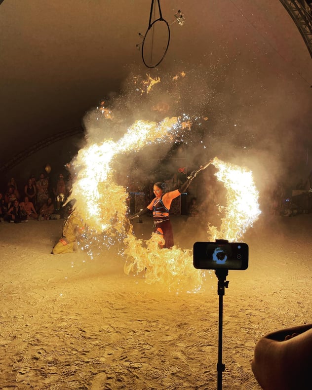 A woman dancing with flames while she participates in a fire performance. There is a crown of people sitting in the background at night.